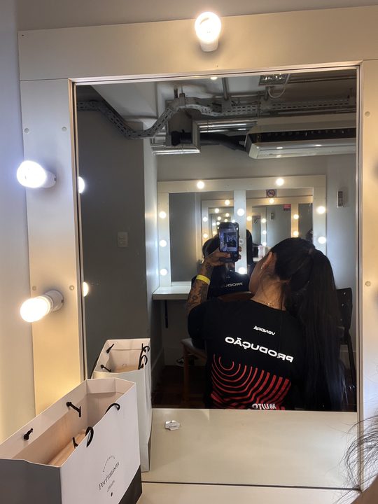 Yasmim de camiseta de produção no camarim, fotografando o espelho
